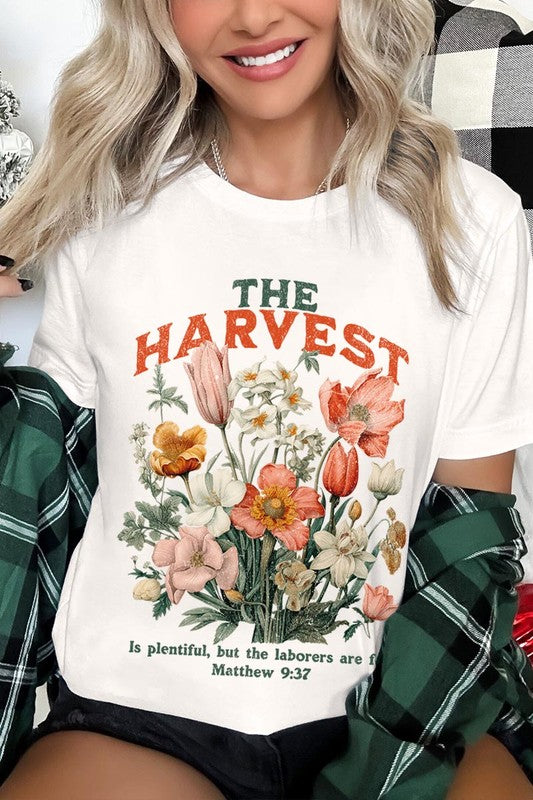 A woman is pictured wearing The Harvest Christian Graphic Tee, which features "The Harvest" in bold letters, an illustration of various flowers, and the verse "Is plentiful, but the laborers are few. Matthew 9:37." She has her denim jacket open to display the tee. This versatile shirt is ideal for Family Group Uniforms or as a Unisex Crew Neck option.