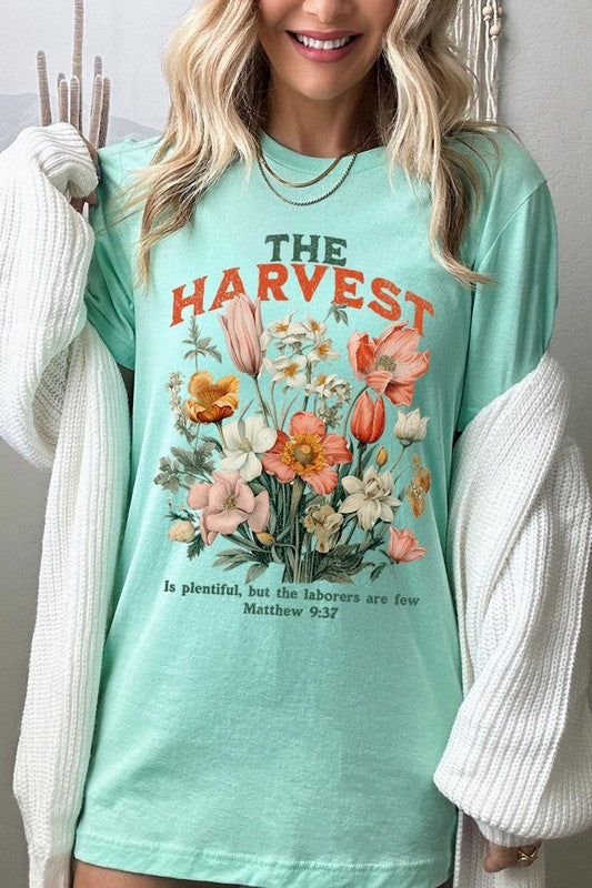 A woman is pictured wearing The Harvest Christian Graphic Tee, which features "The Harvest" in bold letters, an illustration of various flowers, and the verse "Is plentiful, but the laborers are few. Matthew 9:37." She has her denim jacket open to display the tee. This versatile shirt is ideal for Family Group Uniforms or as a Unisex Crew Neck option.