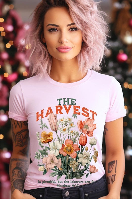 A woman is pictured wearing The Harvest Christian Graphic Tee, which features "The Harvest" in bold letters, an illustration of various flowers, and the verse "Is plentiful, but the laborers are few. Matthew 9:37." She has her denim jacket open to display the tee. This versatile shirt is ideal for Family Group Uniforms or as a Unisex Crew Neck option.