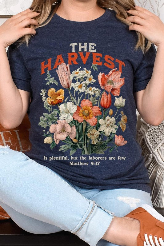 A woman is pictured wearing The Harvest Christian Graphic Tee, which features "The Harvest" in bold letters, an illustration of various flowers, and the verse "Is plentiful, but the laborers are few. Matthew 9:37." She has her denim jacket open to display the tee. This versatile shirt is ideal for Family Group Uniforms or as a Unisex Crew Neck option.