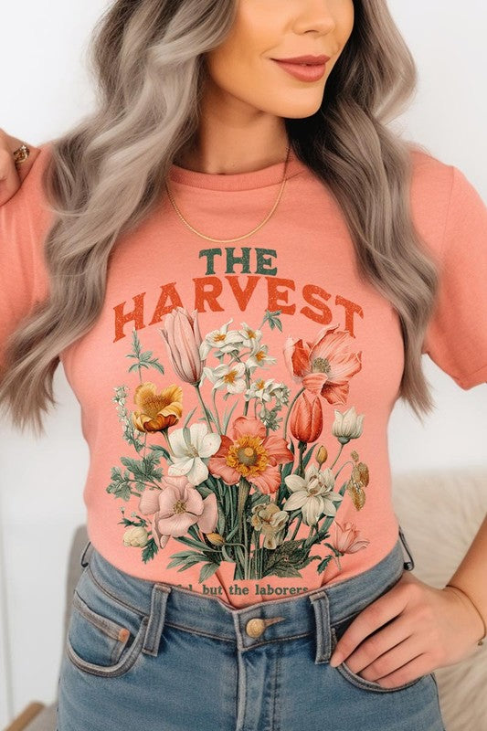 A woman is pictured wearing The Harvest Christian Graphic Tee, which features "The Harvest" in bold letters, an illustration of various flowers, and the verse "Is plentiful, but the laborers are few. Matthew 9:37." She has her denim jacket open to display the tee. This versatile shirt is ideal for Family Group Uniforms or as a Unisex Crew Neck option.