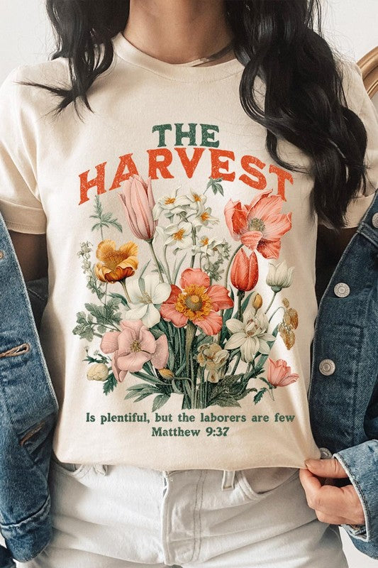 A woman is pictured wearing The Harvest Christian Graphic Tee, which features "The Harvest" in bold letters, an illustration of various flowers, and the verse "Is plentiful, but the laborers are few. Matthew 9:37." She has her denim jacket open to display the tee. This versatile shirt is ideal for Family Group Uniforms or as a Unisex Crew Neck option.