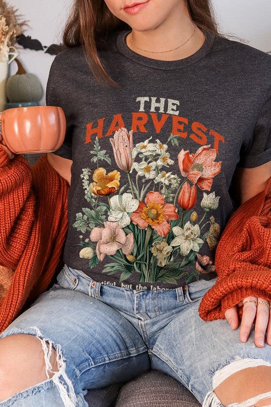 A woman is pictured wearing The Harvest Christian Graphic Tee, which features "The Harvest" in bold letters, an illustration of various flowers, and the verse "Is plentiful, but the laborers are few. Matthew 9:37." She has her denim jacket open to display the tee. This versatile shirt is ideal for Family Group Uniforms or as a Unisex Crew Neck option.