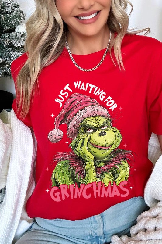 A person with light pink hair stands in front of a festive background, sporting the high-quality Just Waiting For Grinchmas Graphic Tee featuring the Grinch and the text "Just Waiting for Grinchmas.