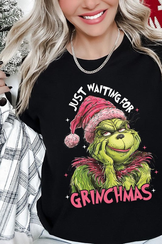 A person with light pink hair stands in front of a festive background, sporting the high-quality Just Waiting For Grinchmas Graphic Tee featuring the Grinch and the text "Just Waiting for Grinchmas.