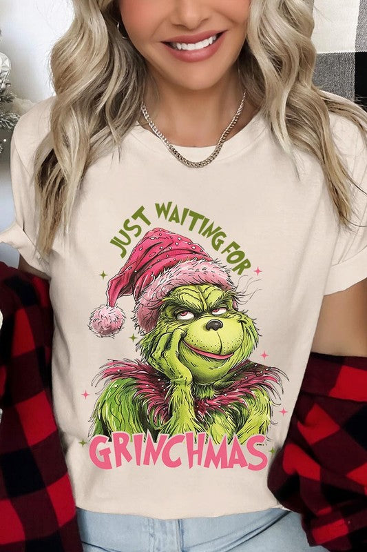 A person with light pink hair stands in front of a festive background, sporting the high-quality Just Waiting For Grinchmas Graphic Tee featuring the Grinch and the text "Just Waiting for Grinchmas.