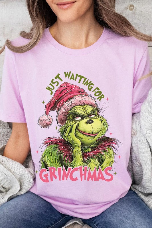 A person with light pink hair stands in front of a festive background, sporting the high-quality Just Waiting For Grinchmas Graphic Tee featuring the Grinch and the text "Just Waiting for Grinchmas.