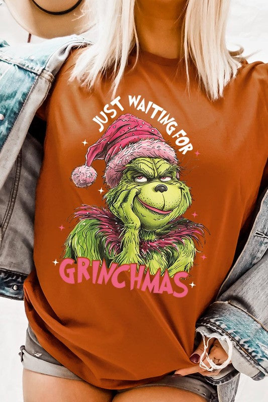 A person with light pink hair stands in front of a festive background, sporting the high-quality Just Waiting For Grinchmas Graphic Tee featuring the Grinch and the text "Just Waiting for Grinchmas.