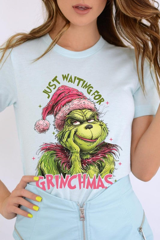 A person with light pink hair stands in front of a festive background, sporting the high-quality Just Waiting For Grinchmas Graphic Tee featuring the Grinch and the text "Just Waiting for Grinchmas.