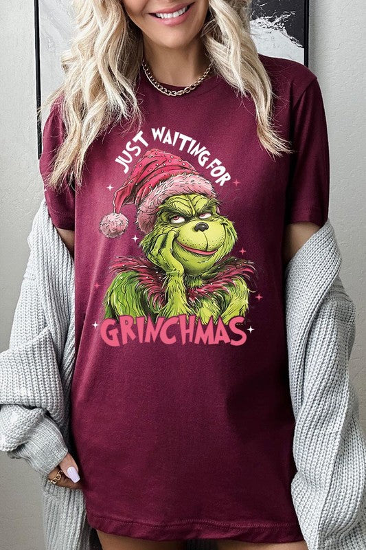 A person with light pink hair stands in front of a festive background, sporting the high-quality Just Waiting For Grinchmas Graphic Tee featuring the Grinch and the text "Just Waiting for Grinchmas.