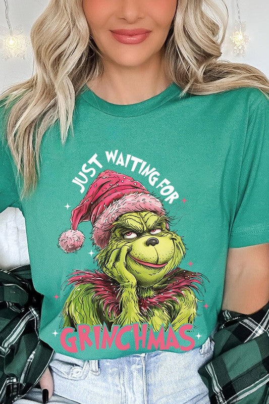 A person with light pink hair stands in front of a festive background, sporting the high-quality Just Waiting For Grinchmas Graphic Tee featuring the Grinch and the text "Just Waiting for Grinchmas.