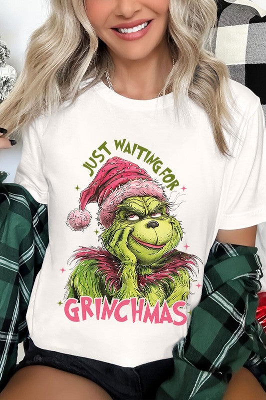 A person with light pink hair stands in front of a festive background, sporting the high-quality Just Waiting For Grinchmas Graphic Tee featuring the Grinch and the text "Just Waiting for Grinchmas.