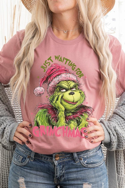 A person with light pink hair stands in front of a festive background, sporting the high-quality Just Waiting For Grinchmas Graphic Tee featuring the Grinch and the text "Just Waiting for Grinchmas.