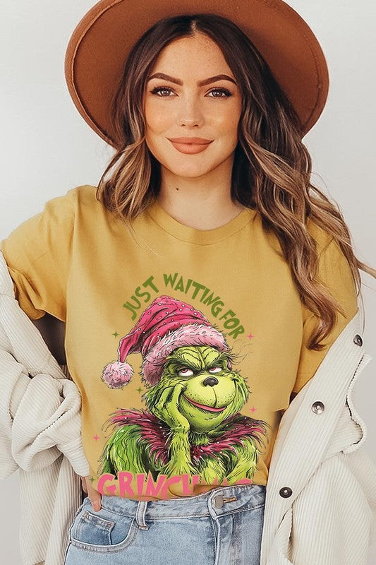 A person with light pink hair stands in front of a festive background, sporting the high-quality Just Waiting For Grinchmas Graphic Tee featuring the Grinch and the text "Just Waiting for Grinchmas.