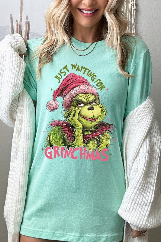 A person with light pink hair stands in front of a festive background, sporting the high-quality Just Waiting For Grinchmas Graphic Tee featuring the Grinch and the text "Just Waiting for Grinchmas.