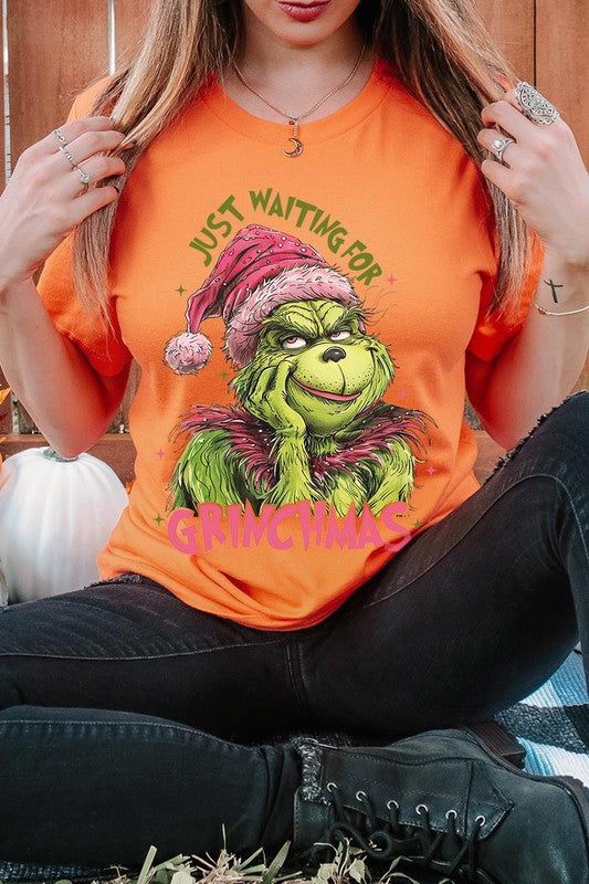 A person with light pink hair stands in front of a festive background, sporting the high-quality Just Waiting For Grinchmas Graphic Tee featuring the Grinch and the text "Just Waiting for Grinchmas.