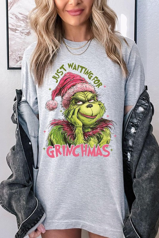 A person with light pink hair stands in front of a festive background, sporting the high-quality Just Waiting For Grinchmas Graphic Tee featuring the Grinch and the text "Just Waiting for Grinchmas.