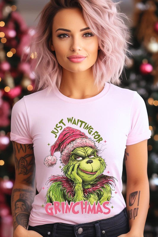 A person with light pink hair stands in front of a festive background, sporting the high-quality Just Waiting For Grinchmas Graphic Tee featuring the Grinch and the text "Just Waiting for Grinchmas.