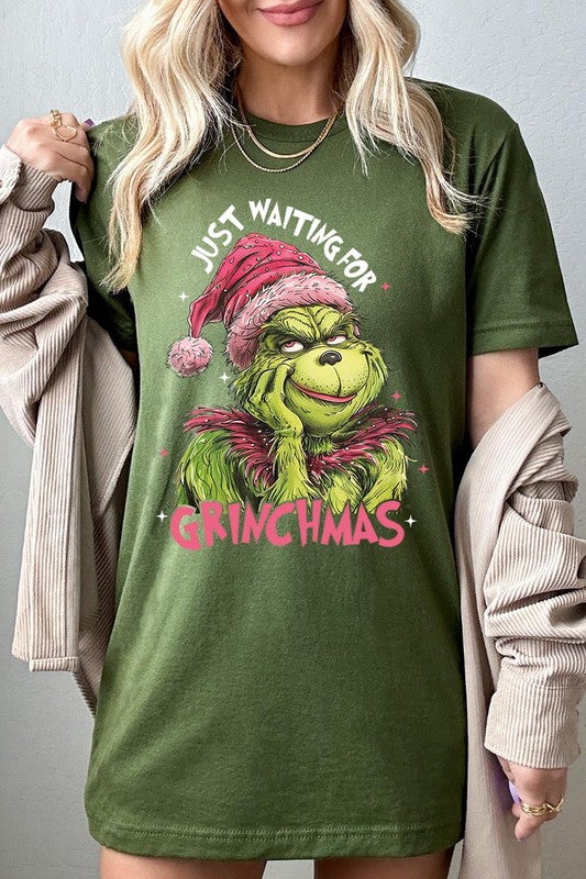 A person with light pink hair stands in front of a festive background, sporting the high-quality Just Waiting For Grinchmas Graphic Tee featuring the Grinch and the text "Just Waiting for Grinchmas.