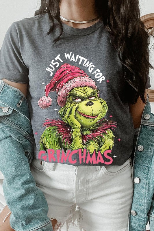 A person with light pink hair stands in front of a festive background, sporting the high-quality Just Waiting For Grinchmas Graphic Tee featuring the Grinch and the text "Just Waiting for Grinchmas.