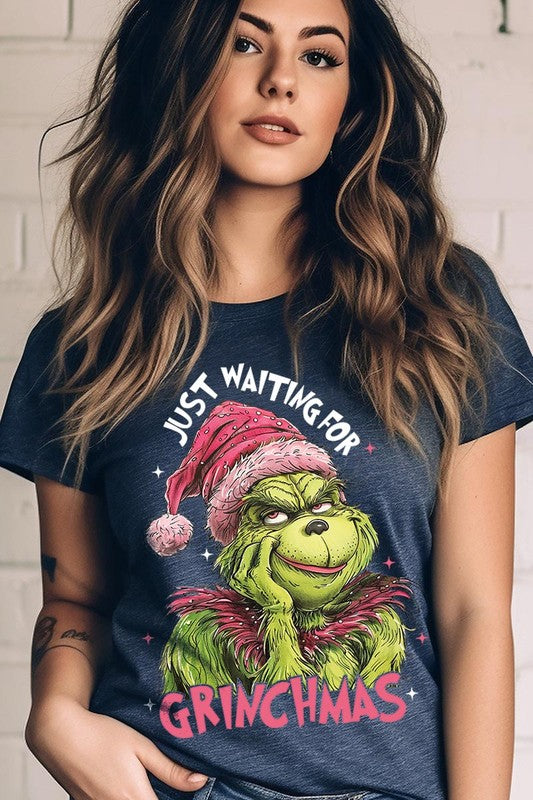 A person with light pink hair stands in front of a festive background, sporting the high-quality Just Waiting For Grinchmas Graphic Tee featuring the Grinch and the text "Just Waiting for Grinchmas.