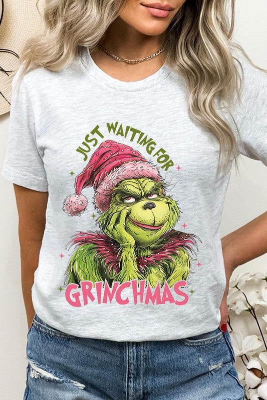 A person with light pink hair stands in front of a festive background, sporting the high-quality Just Waiting For Grinchmas Graphic Tee featuring the Grinch and the text "Just Waiting for Grinchmas.