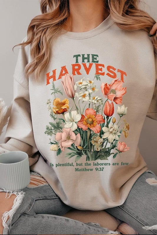 A person wearing 'The Harvest Christian Graphic Fleece Sweatshirt' in navy blue, adorned with floral graphics and the text "The Harvest" alongside "Matthew 9:37 - The harvest is plentiful, but the laborers are few," effortlessly combines style and comfort in this high-quality unisex crew neck.