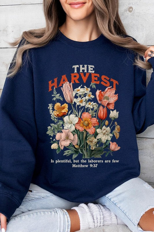 A person wearing 'The Harvest Christian Graphic Fleece Sweatshirt' in navy blue, adorned with floral graphics and the text "The Harvest" alongside "Matthew 9:37 - The harvest is plentiful, but the laborers are few," effortlessly combines style and comfort in this high-quality unisex crew neck.