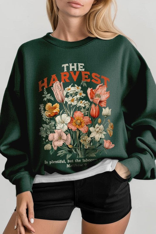 A person wearing 'The Harvest Christian Graphic Fleece Sweatshirt' in navy blue, adorned with floral graphics and the text "The Harvest" alongside "Matthew 9:37 - The harvest is plentiful, but the laborers are few," effortlessly combines style and comfort in this high-quality unisex crew neck.