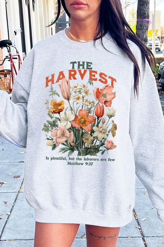 A person wearing 'The Harvest Christian Graphic Fleece Sweatshirt' in navy blue, adorned with floral graphics and the text "The Harvest" alongside "Matthew 9:37 - The harvest is plentiful, but the laborers are few," effortlessly combines style and comfort in this high-quality unisex crew neck.