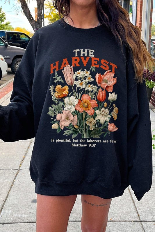 A person wearing 'The Harvest Christian Graphic Fleece Sweatshirt' in navy blue, adorned with floral graphics and the text "The Harvest" alongside "Matthew 9:37 - The harvest is plentiful, but the laborers are few," effortlessly combines style and comfort in this high-quality unisex crew neck.