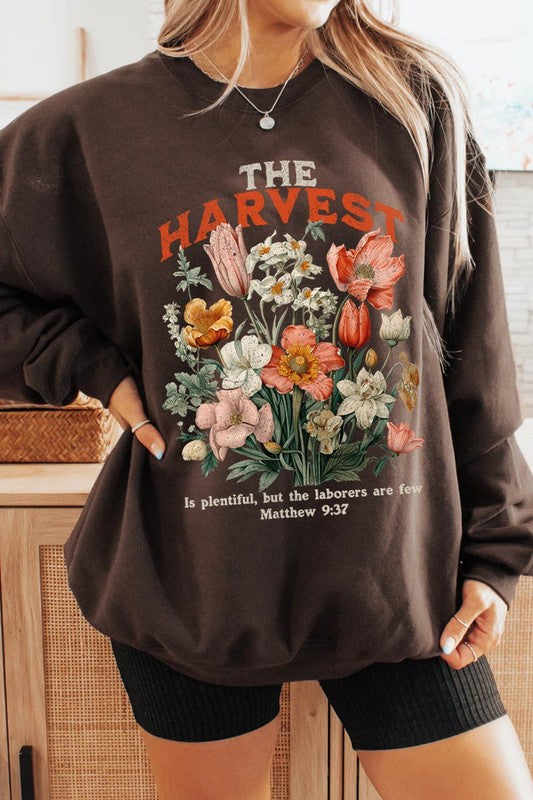 A person wearing 'The Harvest Christian Graphic Fleece Sweatshirt' in navy blue, adorned with floral graphics and the text "The Harvest" alongside "Matthew 9:37 - The harvest is plentiful, but the laborers are few," effortlessly combines style and comfort in this high-quality unisex crew neck.