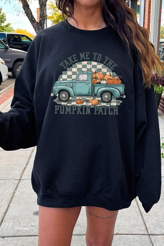 A woman with blonde hair wearing a "Take Me To The Pumpkin Patch" Fleece Sweatshirt stands in front of a vintage truck filled with pumpkins, holding a small device in her right hand.