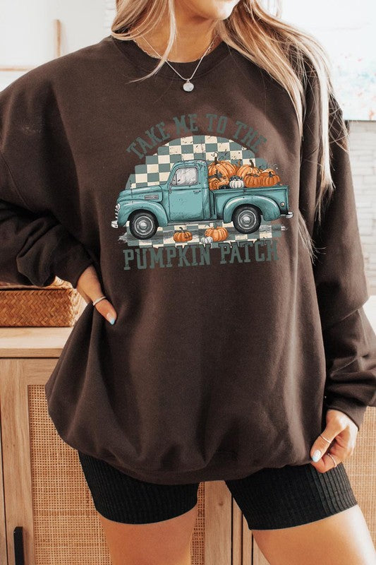 A woman with blonde hair wearing a "Take Me To The Pumpkin Patch" Fleece Sweatshirt stands in front of a vintage truck filled with pumpkins, holding a small device in her right hand.