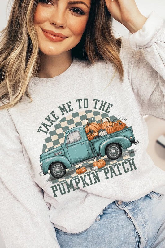 A woman with blonde hair wearing a "Take Me To The Pumpkin Patch" Fleece Sweatshirt stands in front of a vintage truck filled with pumpkins, holding a small device in her right hand.