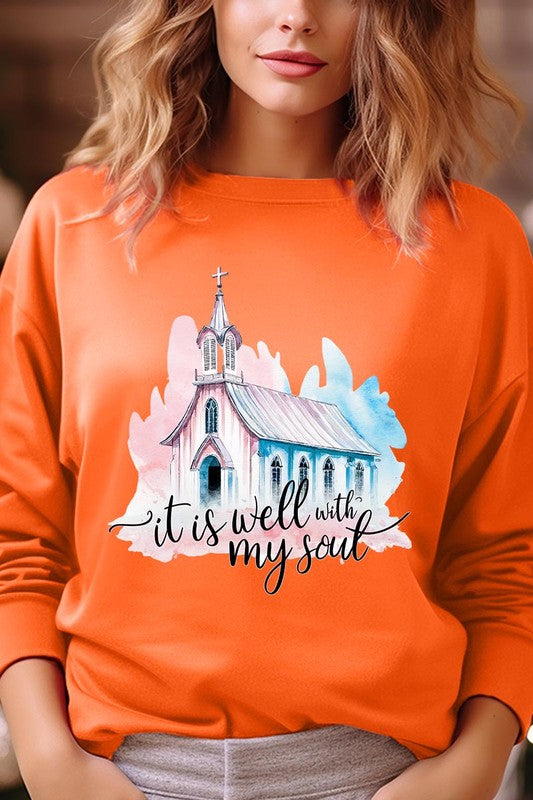 An individual is wearing a bright pink unisex "It Is Well With My Soul Graphic Fleece Sweatshirt," featuring a design of a church and the text "It is well with my soul." The person is holding the collar of the sweatshirt, with a striped blanket visible in the background.