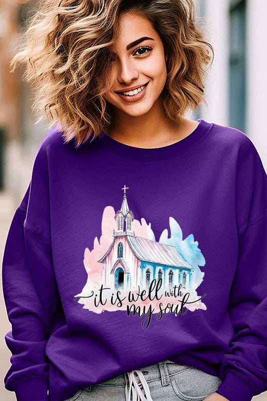 An individual is wearing a bright pink unisex "It Is Well With My Soul Graphic Fleece Sweatshirt," featuring a design of a church and the text "It is well with my soul." The person is holding the collar of the sweatshirt, with a striped blanket visible in the background.