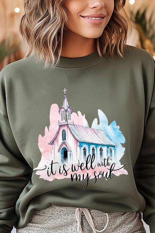 An individual is wearing a bright pink unisex "It Is Well With My Soul Graphic Fleece Sweatshirt," featuring a design of a church and the text "It is well with my soul." The person is holding the collar of the sweatshirt, with a striped blanket visible in the background.