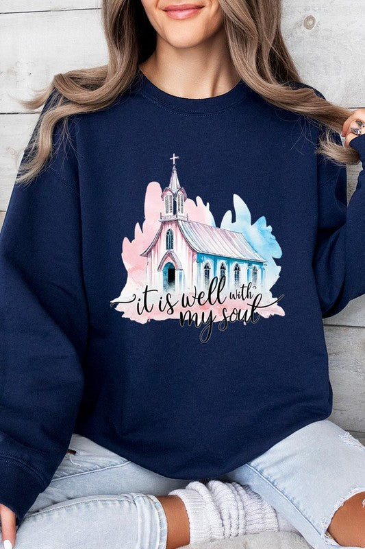 An individual is wearing a bright pink unisex "It Is Well With My Soul Graphic Fleece Sweatshirt," featuring a design of a church and the text "It is well with my soul." The person is holding the collar of the sweatshirt, with a striped blanket visible in the background.