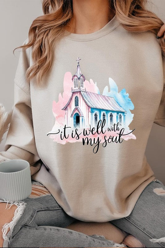 An individual is wearing a bright pink unisex "It Is Well With My Soul Graphic Fleece Sweatshirt," featuring a design of a church and the text "It is well with my soul." The person is holding the collar of the sweatshirt, with a striped blanket visible in the background.