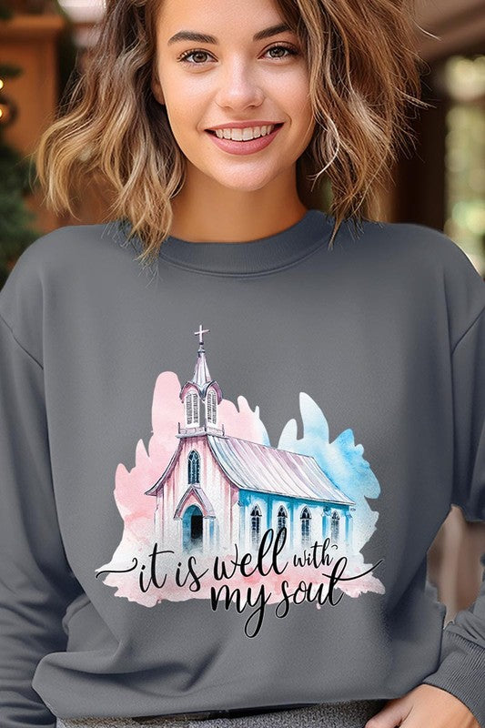 An individual is wearing a bright pink unisex "It Is Well With My Soul Graphic Fleece Sweatshirt," featuring a design of a church and the text "It is well with my soul." The person is holding the collar of the sweatshirt, with a striped blanket visible in the background.