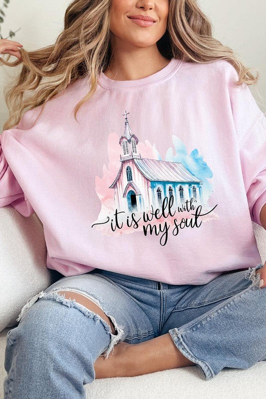 An individual is wearing a bright pink unisex "It Is Well With My Soul Graphic Fleece Sweatshirt," featuring a design of a church and the text "It is well with my soul." The person is holding the collar of the sweatshirt, with a striped blanket visible in the background.