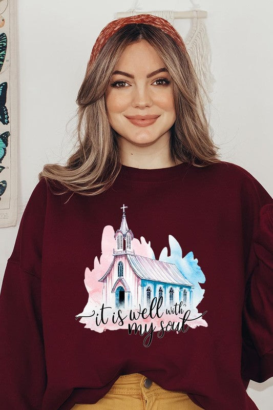 An individual is wearing a bright pink unisex "It Is Well With My Soul Graphic Fleece Sweatshirt," featuring a design of a church and the text "It is well with my soul." The person is holding the collar of the sweatshirt, with a striped blanket visible in the background.