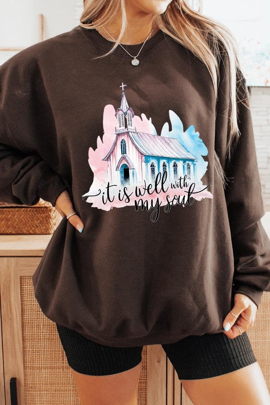 An individual is wearing a bright pink unisex "It Is Well With My Soul Graphic Fleece Sweatshirt," featuring a design of a church and the text "It is well with my soul." The person is holding the collar of the sweatshirt, with a striped blanket visible in the background.