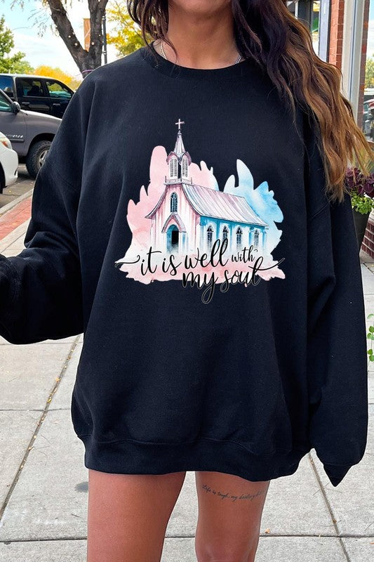 An individual is wearing a bright pink unisex "It Is Well With My Soul Graphic Fleece Sweatshirt," featuring a design of a church and the text "It is well with my soul." The person is holding the collar of the sweatshirt, with a striped blanket visible in the background.