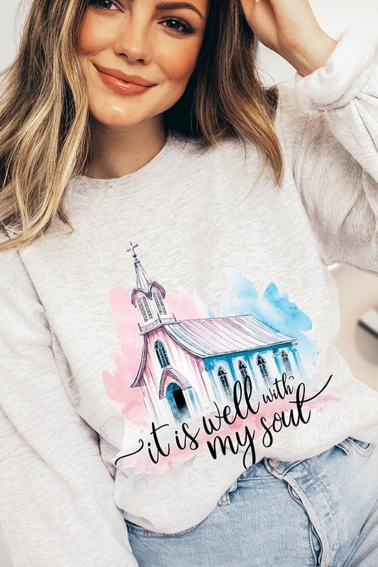 An individual is wearing a bright pink unisex "It Is Well With My Soul Graphic Fleece Sweatshirt," featuring a design of a church and the text "It is well with my soul." The person is holding the collar of the sweatshirt, with a striped blanket visible in the background.