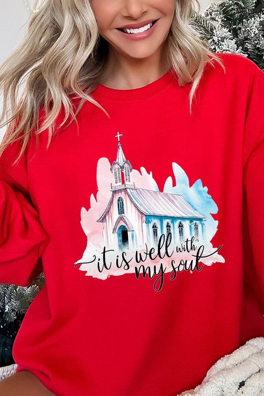 An individual is wearing a bright pink unisex "It Is Well With My Soul Graphic Fleece Sweatshirt," featuring a design of a church and the text "It is well with my soul." The person is holding the collar of the sweatshirt, with a striped blanket visible in the background.