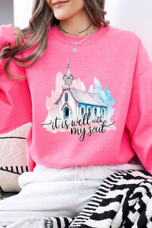 An individual is wearing a bright pink unisex "It Is Well With My Soul Graphic Fleece Sweatshirt," featuring a design of a church and the text "It is well with my soul." The person is holding the collar of the sweatshirt, with a striped blanket visible in the background.