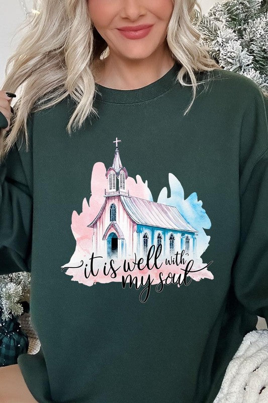 An individual is wearing a bright pink unisex "It Is Well With My Soul Graphic Fleece Sweatshirt," featuring a design of a church and the text "It is well with my soul." The person is holding the collar of the sweatshirt, with a striped blanket visible in the background.