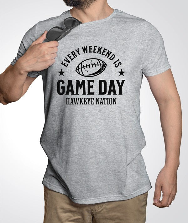 A person stands in a stadium wearing a Gameday Hawkeye Nation Graphic Crew Tee that reads "Every Weekend is Game Day - Hawkeye Nation" with a football graphic. The background includes stadium seating and a grass field.